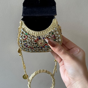 May include: A gold-colored, embellished handbag with a black interior. The bag is adorned with colorful gemstones and has a circular handle. A gold chain with small coin-like charms is attached.
