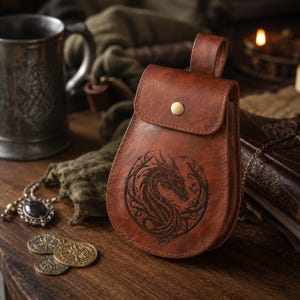 Pode incluir: Uma bolsa de couro castanho com um design de dragão circular em relevo na aba frontal. A bolsa possui um passador de cinto no topo, um fecho de botão de pressão cor de latão e uma forma arredondada estruturada com costuras visíveis.
