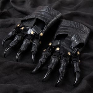 Puede incluir: Un par de guantes negros con un patrón de piel de cocodrilo texturizado en los puños, acentuados con remaches metálicos dorados. Cada sección de los dedos está recubierta de un material negro texturizado y termina en una garra negra larga y curva.
