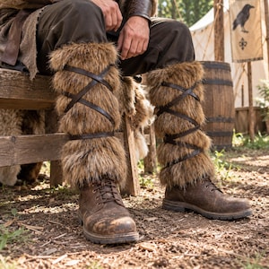 May include: A pair of brown faux fur leg warmers featuring dark brown leather straps that crisscross around the calves. The leg warmers are worn over brown leather lace-up boots, providing a textured, layered appearance for the lower legs.
