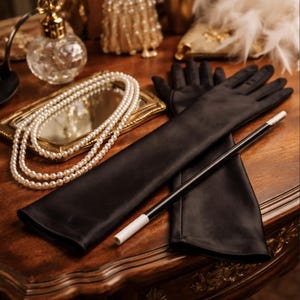 May include: A still-life image featuring a pair of long, black gloves, a magic wand, and a pearl necklace on a wooden surface. The gloves are sleek and elegant, and the wand has a white tip. A small, gold-framed mirror and a perfume bottle are also visible.