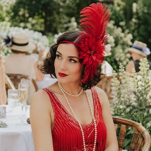 May include: A red feather fascinator headband featuring a large fabric flower and upright plumes. The accessory is paired with a sleeveless red dress with fringe detailing and a long, multi-strand white pearl necklace worn around the neck.