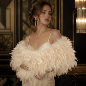 Può includere: Una donna indossa una boa di piume color crema sopra un abito sottoveste di raso. La boa è voluminosa e soffice, con un aspetto morbido e strutturato. Indossa anche gioielli delicati, tra cui una collana e orecchini. Lo sfondo presenta un interno scuro e ornato.