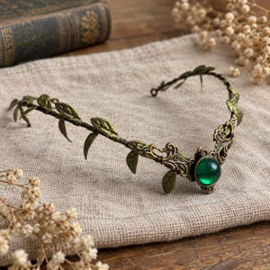 May include: A bronze-toned metal headband featuring a vine-like design with small leaves and a central circular emerald green cabochon stone. The band is constructed with intricate filigree metalwork surrounding the green stone at the center.