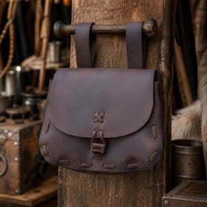 May include: A dark brown leather pouch with a flap closure and stitched details. The pouch hangs from a wooden dowel and leather straps, attached to a wooden post. The design evokes a medieval or historical aesthetic.