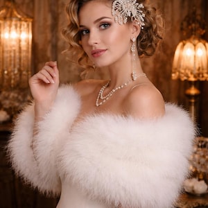 May include: A woman wearing a white feathered shawl, pearl necklace, and jeweled hair accessory. The image is a portrait with a soft focus, highlighting the elegant details of the bridal or formal wear. The background features warm lighting.