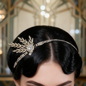 May include: A gold-toned headpiece with a central, large, clear gemstone and smaller surrounding crystals. The headpiece features a leaf-like design on one side and a delicate band of crystals across the forehead. The hair is styled in a classic, dark wave.