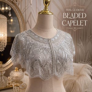 May include: A silver beaded capelet featuring intricate sequin and pearl embellishments on a sheer fabric base. The garment displays a scalloped hemline and a central vertical row of pearls. Text on the image reads 1920s GLAMOUR BEADED CAPELET.
