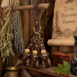 May include: A dark brown leather strap hanger featuring three brass swivel clips, each holding a small clear glass bottle with a cork stopper. The leather straps are secured with brass rivets and hang from a central brass ring and leather loop.