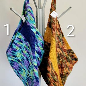 May include: Two handmade crochet tote bags. One bag is primarily blue with green and purple accents. The other bag is yellow, orange, and brown. Both bags are hanging on a silver coat rack. The bags are made of yarn.
