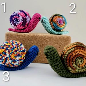 May include: Four handmade crochet snails in various colors. The snails have colorful shells and bodies. One snail has a pink body and a shell with purple, pink, and white stripes. Another has a blue body and a shell with orange, yellow, and blue speckles.