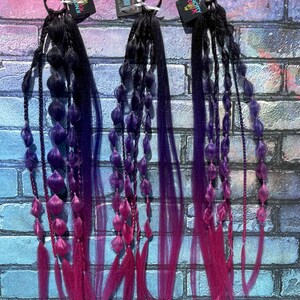 May include: Three hair extensions with black, purple, and pink ombre coloring. Each extension features a black elastic band, braided sections, and decorative beads. The extensions are displayed against a brick wall.