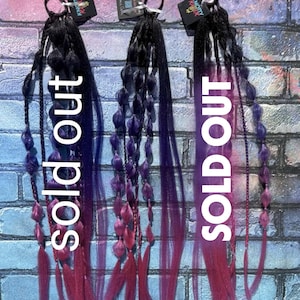 May include: Three hair extensions with black, purple, and pink ombre coloring. Each extension is attached to a black hair tie and has a tag that reads "Knotty Kinky". The word "SOLD OUT" is printed vertically on each extension.
