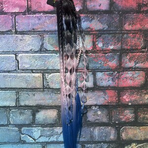 May include: Braided hair extension with a gradient effect, transitioning from black to gray, then to pink, and finally to blue. The extension features decorative beads and a black elastic band for attachment. A small tag is attached to the top.