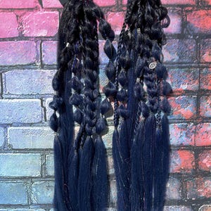 May include: Two dark blue braided hair extensions with a mix of textures, including braids and loose strands. The extensions are attached to black hair ties and adorned with decorative accents. The background is a brick wall with a colorful graffiti design.