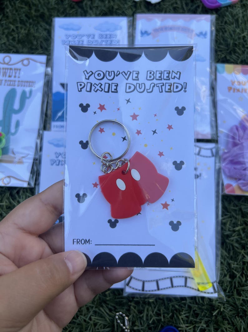 May include: A red Mickey Mouse-shaped keychain with white accents, attached to a silver keyring. The keychain is packaged in a clear plastic bag with a card that reads "YOU'VE BEEN PIXIE DUSTED!" and has Mickey Mouse silhouettes.