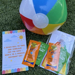 May include: A colorful beach ball, sunscreen packets, and a card with text that reads "A beach ball to toss and sunblock for the rays - here's to a magical castaway day!" on a green surface.