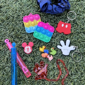 May include: A collection of colorful accessories including a blue scrunchie, two rainbow-colored pop-it keychains, Mickey Mouse-themed keychains, a hand-shaped keychain, toy ducks, bubbles, and a red, hand-shaped toy, all on a green surface.