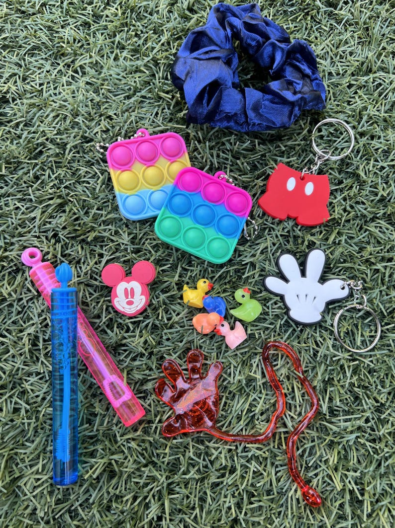 May include: A collection of colorful accessories including a blue scrunchie, two rainbow-colored pop-it keychains, Mickey Mouse-themed keychains, a hand-shaped keychain, toy ducks, bubbles, and a red, hand-shaped toy, all on a green surface.