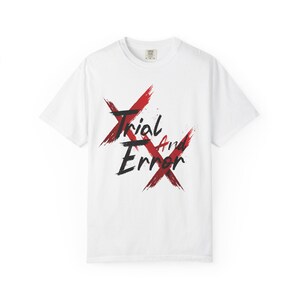 Op de afbeelding: Een wit T-shirt met korte mouwen en een grafisch ontwerp op de borst. Het ontwerp bestaat uit twee grote, elkaar kruisende rode penseelstreek-X-tekens met de zwarte tekst Trial And Error in een gestileerd, handgeschreven lettertype.