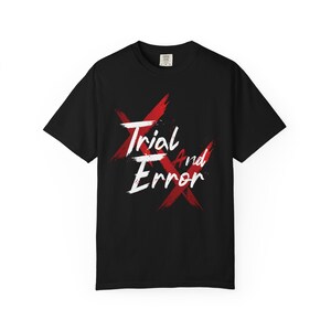 May include: A black short-sleeved t-shirt featuring a graphic design with two large, distressed red X marks layered behind the white text that reads Trial And Error in a stylised, brush-stroke font centred on the front of the garment.