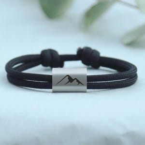 Könnte beinhalten: Ein schwarzes Kordelarmband mit einer zylindrischen, silberfarbenen Metallperle in der Mitte. Die Perle ist mit einer dunklen, minimalistischen Silhouette eines Gebirges graviert. Das Armband verfügt über verstellbare Schiebeknoten.