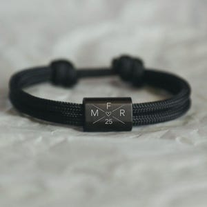 Könnte beinhalten: Schwarz geflochtenes Armband mit rechteckigem Metallakzent. Der Akzent zeigt die Buchstaben "F", "M" und "R", ein Herzsymbol und die Zahl "25". Das Armband ist an jedem Ende geknotet.
