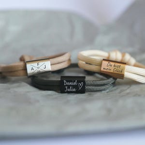 Könnte beinhalten: Drei personalisierte Armbänder mit geflochtenen Kordelbändern. Ein Armband ist beige mit einem silbernen Verschluss, der mit Initialen und einem Datum graviert ist. Ein weiteres ist cremefarben mit einem goldenen Verschluss. Das dritte ist schwarz und grau mit einem schwarzen Verschluss.