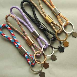 Könnte beinhalten: Sechs Schlüsselanhänger aus geflochtenem Seil mit Metallschließe und hausförmigem Anhänger. Die Seile sind rot-weiß-blau, bunt, lavendel, schwarz, beige und orange-gelb-weiß. Die Metallschließen tragen deutsche Gravuren wie Danke für alles.