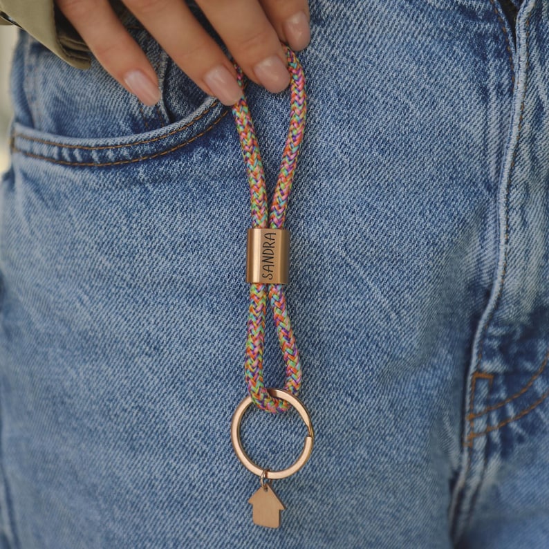 Puede incluir: Un llavero colorido con un cord&oacute;n trenzado en tonos rosa, amarillo, verde y azul. El llavero presenta un anillo de metal dorado y un peque&ntilde;o dije en forma de casa. Tambi&eacute;n hay un tubo de metal dorado con el nombre "SANDRA".