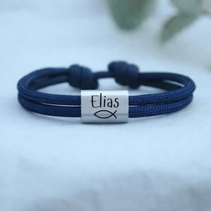 Könnte beinhalten: Ein marineblaues Armband mit einem silbernen, rechteckigen Anhänger. Der Anhänger ist mit dem Namen "Elias" und einem Fischsymbol graviert. Das Armband besteht aus zwei zusammengeknoteten Kordeln.