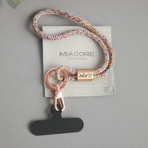 Könnte beinhalten: Ein mehrfarbiges geflochtenes Schlüsselband mit einem roségoldenen Verschluss und einem rechteckigen Anhänger mit der Gravur "Julia ♡". Das Schlüsselband ist an einem schwarzen Handyhalter befestigt und liegt auf einem grauen Beutel mit dem Logo "MIACore Handmade".
