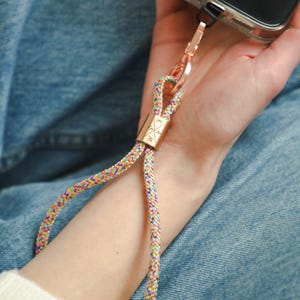 Könnte beinhalten: Eine mehrfarbige geflochtene Handgelenkschlaufe für das Telefon mit einem goldfarbenen Verschluss und einem goldfarbenen rechteckigen Akzentstück. Die Schlaufe ist an einer transparenten Handyhülle befestigt. Der Hintergrund ist eine Person in Jeans.