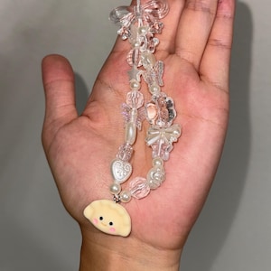 May include: A clear, beaded phone charm with a dumpling charm. The charm features clear and pink beads, pearls, and clear butterfly and bow accents. The dumpling charm is yellow with a smiling face.