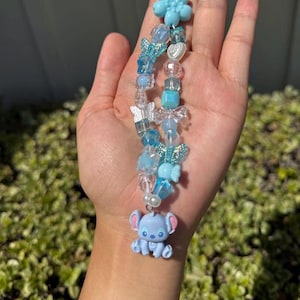 May include: A blue beaded charm featuring a Stitch figurine, butterflies, and flower accents. The charm is strung with various blue and clear beads, with a white string loop at the top. The charm is held in a hand.