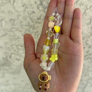 May include: A beaded phone charm featuring a brown bear charm with a yellow hat. The charm is strung with a variety of beads in yellow, white, and clear colors. The charm is attached to a white string.