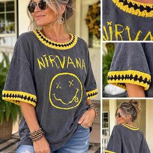 May include: A charcoal gray t-shirt featuring the yellow text NIRVANA and a yellow smiley face graphic. The neckline and sleeve cuffs are finished with a decorative yellow and black crocheted trim. The shirt has a relaxed fit and short sleeves.
