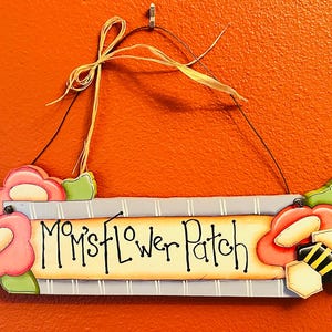 May include: A wooden sign with the text "Mom's Flower Patch" in black script. The sign is decorated with pink and green flowers, and a bee. The sign hangs from a string and is against an orange background.