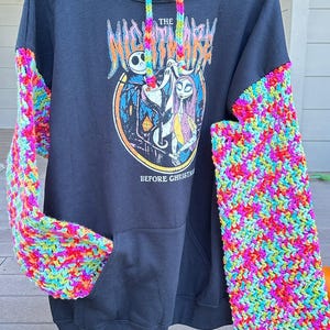 Puede incluir: Sudadera con capucha negra con mangas de ganchillo coloridas. La parte delantera presenta un gr&aacute;fico de "Pesadilla antes de Navidad" con Jack Skellington y Sally. El texto "The Nightmare Before Christmas" y "Before Christmas" es visible.