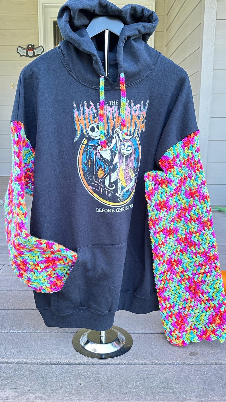 Puede incluir: Sudadera con capucha negra con mangas de ganchillo coloridas. La parte delantera presenta un gr&aacute;fico de "Pesadilla antes de Navidad" con Jack Skellington y Sally. El texto "The Nightmare Before Christmas" y "Before Christmas" es visible.