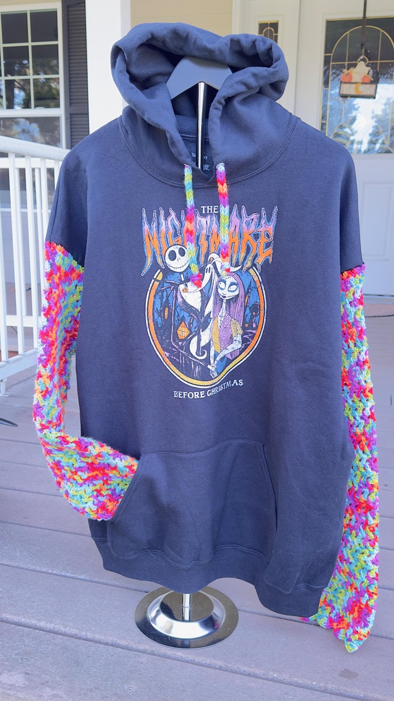 Puede incluir: Sudadera con capucha azul marino con un gr&aacute;fico de "Pesadilla antes de Navidad". Las mangas est&aacute;n tejidas con un patr&oacute;n de arco&iacute;ris. La parte delantera presenta un dise&ntilde;o circular con Jack Skellington y Sally. El texto dice "The Nightmare Before Christmas".