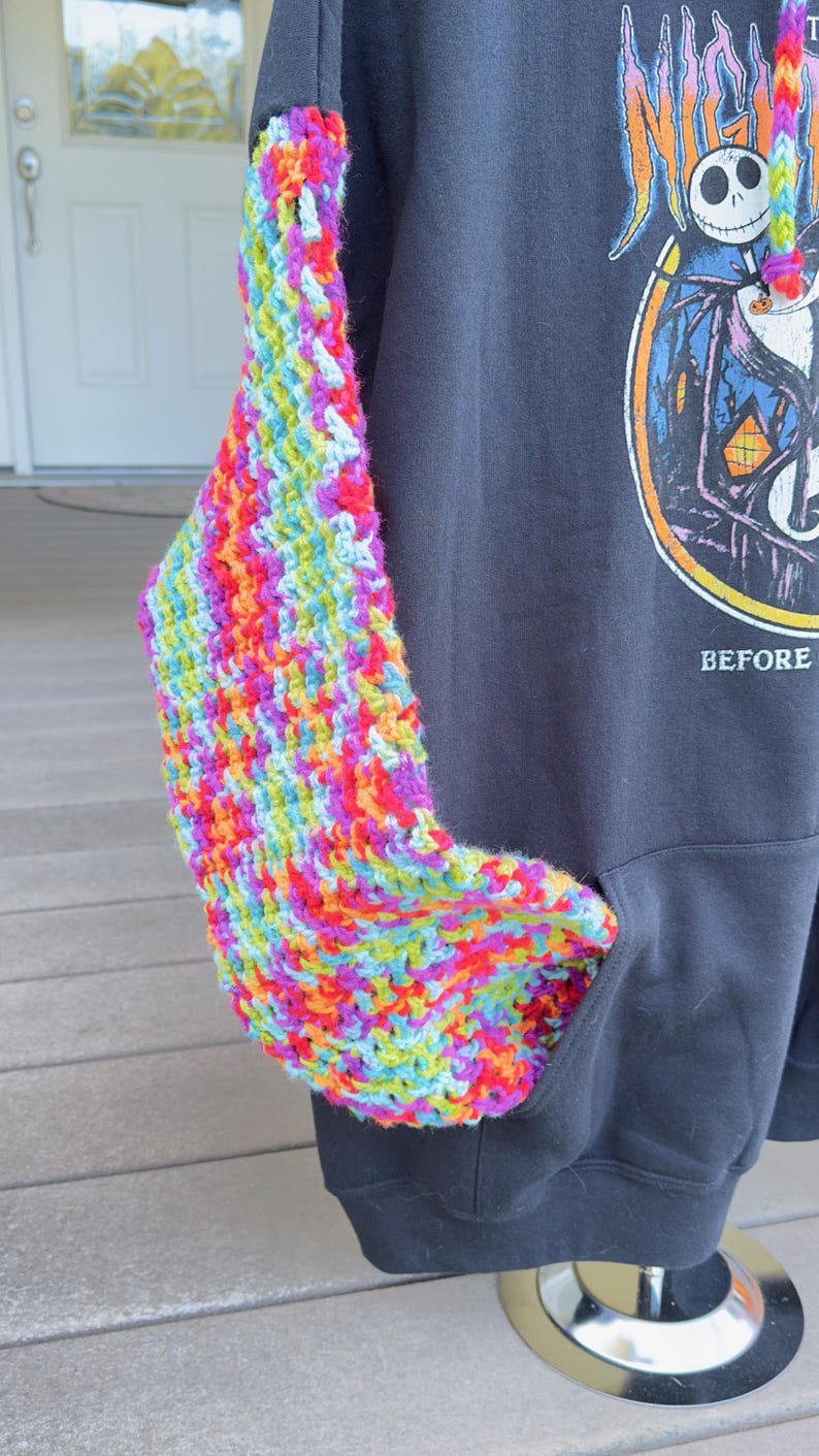 Puede incluir: Una sudadera con capucha negra con una manga y un bolsillo de ganchillo coloridos. La manga est&aacute; hecha de hilo vibrante en tonos de rojo, naranja, amarillo, verde, azul y morado. La sudadera tiene un gr&aacute;fico en la parte delantera.