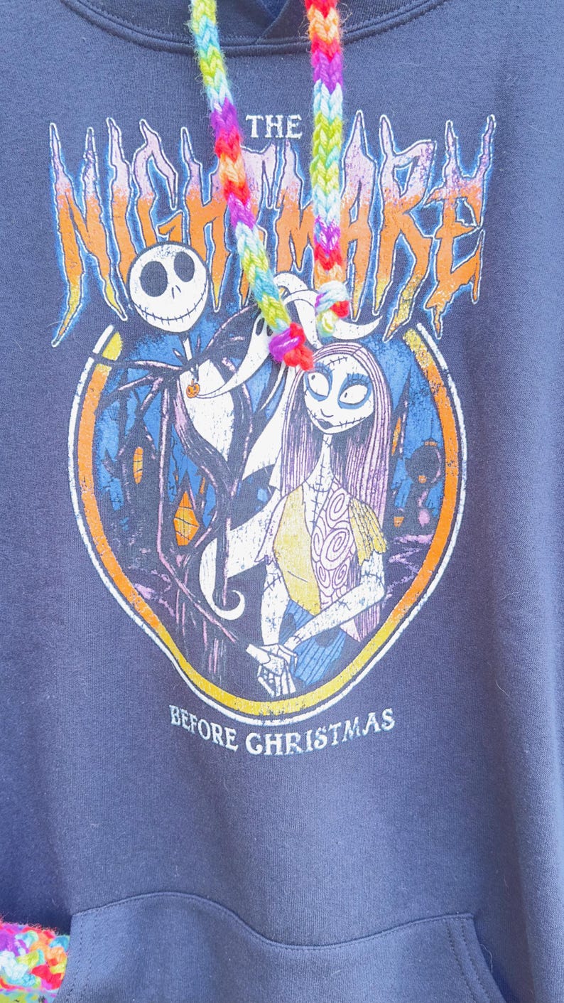 Puede incluir: Sudadera con capucha azul marino con un dise&ntilde;o gr&aacute;fico de "Pesadilla antes de Navidad". El dise&ntilde;o incluye a Jack Skellington y Sally, con el texto "The Nightmare Before Christmas". La capucha tiene un cord&oacute;n arco&iacute;ris.