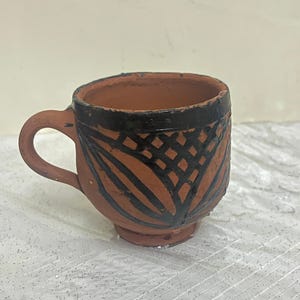 Puede incluir: Una pequeña taza de terracota hecha a mano con asa. La taza presenta un borde marrón oscuro y un patrón decorativo de líneas y formas negras. La taza está sobre una superficie blanca.