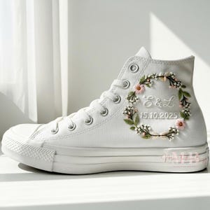 May include: White high-top sneaker with a floral embroidery design. The embroidery includes pink and white flowers, green leaves, and the initials "E&L" with the date "15.10.2025". The shoe has a white rubber sole and laces.
