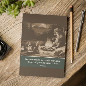 Könnte beinhalten: Ein braunes Notizbuch mit einer Sepia-Illustration von Bären am Lagerfeuer. Es enthält das Zitat "Ich kann niemandem etwas beibringen; ich kann sie nur zum Denken bringen." von Sokrates. Ein Bleistift und ein Stift liegen daneben.