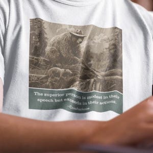 Peut inclure: Deux personnes portent des t-shirts avec un motif sépia d'ours et la citation : « La personne supérieure est modeste dans ses paroles, mais excelle dans ses actions. » Un t-shirt est blanc, l'autre noir.
