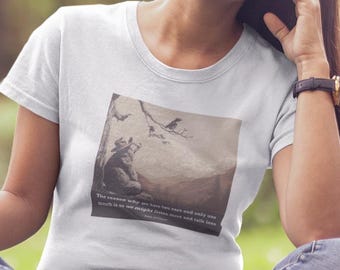 Könnte beinhalten: Weißes T-Shirt mit einem quadratischen Grafikmotiv mit einem Schwarz-Weiß-Bild einer Berglandschaft und Text. Die Person sitzt im Schneidersitz auf dem Gras, trägt eine blaue Jeans und lächelt. Am Handgelenk befindet sich eine schwarze Uhr.