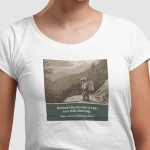 Puede incluir: Camiseta blanca con una imagen en tonos sepia de excursionistas en un paisaje montañoso. La imagen incluye la cita "Detrás de las nubes el sol sigue brillando." de Henry Wadsworth Longfellow.