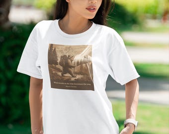 Könnte beinhalten: Weißes T-Shirt mit einem sepiafarbenen Grafik eines Wanderers in einer Berglandschaft. Das Shirt enthält das Zitat: „Was im Weg steht, wird zum Weg“. Das Model trägt eine schwarze Sonnenbrille und blaue Jeans.